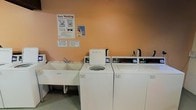 This shared laundry facility is a practical, single-floor space designed to meet the everyday laundering needs of its users. The room features a row of top-loading washing machines equipped with digital controls, allowing for convenient and efficient operation. Each machine is clearly labeled with pricing information, making the process straightforward for all users. Alongside the washers, a large white utility sink with a faucet provides additional functionality for hand-washing or pre-treating garments. The walls are painted in a warm peach tone, lending a modest brightness to the utilitarian space. Instructional posters, including an "Easy Washing" guide and a courtesy notice reminding users to start washers before leaving, are prominently displayed on the wall, ensuring a smooth and considerate experience for all. The flooring is durable and easy to maintain, suited to the demands of a high-traffic laundry environment. Overall, the space is straightforward and efficiently laid out, prioritizing practicality and ease of use for shared residential or commercial laundry needs.