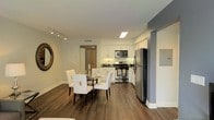 This single-floor residence showcases a thoughtfully designed open-concept layout that seamlessly connects the living, dining, and kitchen areas into one cohesive and functional space. Rich, dark hardwood flooring runs throughout, grounding the interior with warmth and elegance.

The kitchen is well-appointed with stainless steel appliances, including a gas range, built-in microwave, and a full-size refrigerator. Crisp white cabinetry extends to the ceiling, offering generous storage, while dark countertops provide a striking contrast that adds a touch of sophistication. A skylight panel above the kitchen floods the space with natural light, enhancing the bright and airy atmosphere.

The adjacent dining area comfortably accommodates a round glass-top table surrounded by white upholstered chairs, creating an inviting space for meals and entertaining. Track lighting along the ceiling provides adjustable illumination throughout the open area, complementing the natural light.

The overall ambiance is clean, modern, and welcoming, with a neutral color palette of whites and grays accented by the warmth of the hardwood floors. The open sightlines between rooms contribute to a spacious and connected feel, making this residence both practical and visually appealing.
