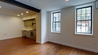 This single-floor space combines classic character with modern updates, creating a warm and functional living environment. Rich hardwood floors run throughout, adding warmth and continuity to the open layout. The main living area is bright and airy, benefiting from large multi-pane windows that draw in generous natural light and offer views of the surrounding brick exterior architecture, lending an urban charm to the interior.

A defining architectural detail is the exposed dark wood beam that frames the transition between the living area and the kitchen, adding a distinctive industrial-rustic touch to the otherwise clean, white-walled space. Recessed lighting is installed throughout, providing flexible and even illumination.

The kitchen is a well-appointed, efficient space featuring stainless steel appliances including a range with oven and a dishwasher, complemented by sleek cabinetry and a practical countertop layout. A utility sink is neatly integrated into the cabinetry, making the kitchen both stylish and highly functional.

Overall, the space strikes a balance between historic character — reflected in the hardwood floors and exposed beam — and contemporary finishes, making it a compelling and comfortable environment suited for everyday living.