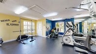 Located in Saint Petersburg, FL, this fitness center offers a well-equipped workout space designed to support a comprehensive exercise routine. The room features a striking two-tone color scheme, with bright yellow walls on three sides and a bold blue accent wall, creating an energetic and motivating atmosphere. Inspirational phrases such as "Never Give Up" and "Be Stronger Than Your Excuses" are displayed prominently on the walls, reinforcing the space's purpose.

The flooring consists of durable dark rubber tile, ideal for both heavy equipment and high-intensity workouts. Overhead, fluorescent lighting fixtures provide consistent, even illumination throughout, complemented by natural light streaming in from multiple windows that offer views of the surrounding Florida landscape.

The strength training area is well-appointed with Hoist-brand cable machines, a power rack with a weight bench, a free-weight station with a barbell and plate storage, and additional selectorized machines for targeted muscle group training. The cardio section includes an elliptical trainer and a stationary bike, accommodating a variety of aerobic workouts. A wall-mounted mirror on one side enhances the sense of space and allows users to monitor their form. The room also includes a water cooler for convenience and a fire extinguisher for safety compliance.
