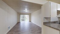 Located at 16124 SE Alder St in Portland, Oregon, this single-floor unit offers a practical and well-appointed layout designed for comfortable everyday living. The living area is bright and open, featuring light wood-style flooring throughout and a large sliding glass door fitted with vertical blinds that allows natural light to fill the room while providing access to the outdoors. The neutral-toned walls and textured ceilings contribute to a clean, understated aesthetic.

The galley-style kitchen is efficiently designed, with white cabinetry lining both sides, granite-look countertops, a tile backsplash, and a full suite of stainless steel appliances including a refrigerator and dishwasher. A utility panel is conveniently accessible near the kitchen entrance. The kitchen connects directly to the rest of the unit, creating a functional flow between spaces.

The bathroom is neatly finished with a granite-topped vanity, cream cabinetry, a wall-mounted towel bar, and tile wainscoting around the toilet area. Light wood-style flooring continues here, maintaining visual consistency throughout the unit. Additional features include wall-mounted heating units in the living space and a paneled interior door leading between rooms. The overall layout, as reflected in the dollhouse view, is compact yet well-organized, making efficient use of every square foot.