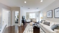 This single-floor residence exudes a clean, contemporary aesthetic rooted in timeless design. The living area serves as the heart of the home, bathed in natural light streaming through white-shuttered windows and complemented by recessed ceiling lighting that creates a warm, welcoming ambiance throughout the day and evening. Smooth, light-toned walls provide a neutral canvas that enhances the sense of openness and space.

Rich, dark hardwood floors run throughout, grounding the room with warmth and elegance. The living space is thoughtfully arranged, featuring a generous white sofa adorned with patterned throw pillows, a dark wood coffee table, and a striking black-and-white geometric area rug that anchors the seating area. A pair of classic white accent chairs and coordinating side tables complete the conversational arrangement, while a dark wood media console adds a bold contrast against the light walls.

Transitioning from the living area, a glimpse into the bedroom reveals a similarly serene palette with layered bedding and natural textures. The open layout and sightlines between rooms contribute to a cohesive and airy feel. Decorative elements — framed artwork, sculptural branches, and accent lamps — are carefully curated to add character without overwhelming the space. Overall, this home offers a refined, comfortable environment with a harmonious balance of light, texture, and neutral sophistication.