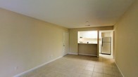 This single-floor space features an open and functional layout with a seamless connection between the living area and kitchen. The main living space is finished with light-toned ceramic tile flooring throughout, contributing to a clean and cohesive aesthetic. Neutral beige walls complement the bright, airy atmosphere of the interior.

The kitchen is accessible via a pass-through counter cutout, which opens directly to the living area and enhances the sense of openness. White cabinetry lines the upper portion of the kitchen, providing ample storage, while a full-size refrigerator is visible along the far wall. The compact kitchen layout is well-organized and practical for everyday use.

A white interior door is positioned along one wall of the main living area, providing access to an adjoining room or closet space. White baseboards run throughout, offering a crisp contrast against the tile floors and warm wall tones. Recessed lighting in the ceiling provides overhead illumination, while the overall layout promotes an efficient and comfortable use of space throughout the unit.