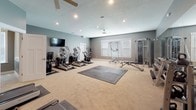 This dedicated fitness center offers a comprehensive workout environment designed to accommodate a wide range of exercise needs. The space is finished with neutral carpet flooring, complemented by cool grey walls and white trim, creating a clean and focused atmosphere throughout.

The main workout area is generously sized, providing ample room for both cardio and strength training. Cardio equipment includes multiple treadmills, elliptical machines, and a stationary spin bike, each positioned on protective rubber mats. A wall-mounted flat-screen television is positioned for easy viewing during workouts. A ceiling fan with integrated lighting ensures comfortable airflow and illumination.

The strength training section features a cable machine and multi-functional weight rack, accompanied by an adjustable workout bench and a medicine ball rack stocked with various weighted balls. A central rubber mat area offers open floor space for stretching, bodyweight exercises, or functional training. A full-length mirror along one wall enhances spatial awareness during workouts.

Natural light filters in through windows fitted with horizontal blinds, and recessed lighting throughout provides consistent illumination. The space also includes a dedicated side room, accessible via a doorway, which offers additional functional space. Emergency exit signage and a fire extinguisher reflect the facility's attention to safety standards.