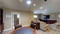 The theater room at 1617 Live Oak in Vista is a thoughtfully designed entertainment space that seamlessly blends a media viewing area with a game room section. The room features warm taupe walls, recessed lighting, and carpet flooring throughout, creating a comfortable and inviting atmosphere.

The primary viewing area is anchored by a large wall-mounted flat-screen television positioned above a rich dark-wood media console with ample cabinet storage. Multiple windows along the walls allow natural light to filter in. Seating is generously arranged, including a full sofa, a loveseat, and several patterned accent chairs surrounding a glass-top coffee table — providing comfortable accommodations for a group. A wall-mounted mini-split HVAC unit ensures independent climate control for the space.

Toward one end of the room, a dedicated game area features a round poker/game table with leather-upholstered chairs, ideal for card games or casual gatherings. A wooden bookcase provides additional storage nearby. A glass-paned door separates this area, offering a degree of privacy while maintaining visual connectivity.

The overall layout is open and functional, making the space suitable for a variety of entertainment and social activities.