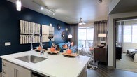 This single-floor space showcases an open-concept layout that integrates the kitchen and living area into one cohesive, stylish environment. The kitchen features a large island with a sleek white quartz countertop, an undermount sink with a contemporary high-arc faucet, and stainless steel appliances. The island serves as both a functional workspace and a natural dividing point between the kitchen and living area.

The living space is anchored by a deep navy blue accent wall that creates a dramatic backdrop for the carefully curated décor, including decorative sunburst mirrors and abstract wall art. Dark hardwood-style flooring runs throughout, complementing the rich color palette. A ceiling fan with integrated lighting occupies the center of the room, while a track lighting system in the kitchen provides focused illumination.

Large windows dressed with floor-to-ceiling gray curtains allow abundant natural light to pour in and offer views of an outdoor area. The overall ambiance blends bold, contemporary design with warm, inviting accents, creating a space that is both visually striking and functionally practical.