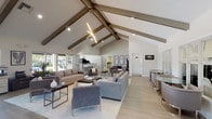 This single-floor community clubhouse is defined by its striking architectural design, centered around soaring vaulted ceilings adorned with distinctive exposed wood beams that span the full length of the open-plan interior. A sleek, angular pendant light fixture runs along the ceiling's peak, providing both functional illumination and a bold design statement.

The main lounge area is generously proportioned, featuring multiple seating arrangements with contemporary gray sofas, armchairs, and glass-topped coffee tables arranged atop light area rugs. The light-toned hardwood flooring throughout enhances the bright, airy atmosphere created by an abundance of natural light streaming through large windows and expansive sliding glass doors.

A built-in bar counter with white cabinetry, tile accents, and modern bar stools serves as a social hub, positioned adjacent to a professional-grade coffee and beverage station. The sliding glass doors open directly onto an outdoor pool area, seamlessly blending the interior and exterior spaces.

Additional functional areas include a dedicated workspace with desks and office equipment, a media lounge featuring a flat-screen TV flanked by framed wall art, and a private office or meeting room with French doors. An ornate entry door with decorative glass work provides a distinguished point of arrival. The overall ambiance is clean, contemporary, and welcoming.