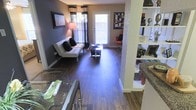 This single-floor residence showcases an open-concept design that connects the dining, living, and kitchen areas into one cohesive, flowing space. Rich, dark hardwood-style flooring runs throughout, providing a warm and stylish foundation that ties the rooms together. A decorative column subtly delineates the dining area from the living room without disrupting the open feel of the layout.

The living area is well-lit, benefiting from a large window and a glass-paneled door that invite abundant natural light, creating a bright and welcoming atmosphere. The neutral wall tones, accented by a bold dark feature wall, add depth and contrast to the space. A shelving unit along one wall offers both display and storage opportunities.

The kitchen, visible from the dining area, features dark granite-style countertops and cabinetry, contributing to the unit's cohesive modern aesthetic. The dining area accommodates a glass-top table, complementing the contemporary styling found throughout. Overall, the space presents a practical yet stylish interior with consistent finishes and a layout designed to maximize functionality and comfort.