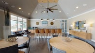 This single-story clubhouse is designed to serve as a welcoming communal gathering space, combining modern aesthetics with functional amenities. The architecture is defined by soaring vaulted ceilings accented by a striking skylight detail, with a large ceiling fan and industrial-style pendant lights anchoring the central gathering area. Polished concrete floors run throughout, reflecting light and lending the space a sleek, contemporary feel.

The main area features multiple round wood dining tables with modern seating, creating a flexible layout for both casual and social gatherings. A full bar counter with a mosaic tile backsplash runs along one wall, lined with leather-upholstered bar stools and equipped with countertop appliances, ideal for events and entertaining. Decorative circular wall accents and curated artwork add visual interest throughout.

An adjacent lounge area offers comfortable seating arranged around a stone-clad fireplace with a wall-mounted television above, complemented by bold yellow accent chairs and a sleek media console. Glass-paned interior doors lead to a separate room, visible as a business or conference space. Large floor-to-ceiling windows and glass doors at the rear open directly to an outdoor pool area, filling the clubhouse with natural light and providing seamless indoor-outdoor connectivity.