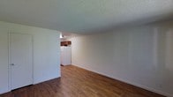 This single-floor space offers a straightforward, functional layout designed for comfortable everyday living. The main living area is characterized by rich, dark hardwood-style flooring that runs throughout, providing a warm and grounding aesthetic. The walls are constructed of painted brick, giving the space a sturdy, solid feel while maintaining a clean, neutral appearance. A textured popcorn ceiling adds depth overhead and helps define the room's modest but practical proportions. The open layout flows directly into the kitchen area, which is visible through a wide pass-through opening. The kitchen is equipped with warm-toned wood cabinetry mounted above the counter, offering ample storage, and includes a full-size white refrigerator. The ceiling-mounted light fixture in the kitchen provides focused illumination to the cooking space. White baseboard trim lines the perimeter of the living area, offering a crisp contrast against the painted brick walls and dark flooring. The overall ambiance is clean and utilitarian, with a neutral color palette throughout that makes the space adaptable to a variety of furnishing styles. The single-floor design ensures easy accessibility and a cohesive flow between the living and kitchen areas.