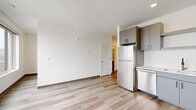 Located at 2132 Stokes Blvd in Cleveland's University Circle neighborhood, this modern single-floor unit offers a well-designed layout encompassing a combined kitchen and living area, one bedroom, two closets, a bathroom, and an in-unit laundry space. Light-toned hardwood-style flooring flows consistently throughout, lending a clean, cohesive aesthetic.

The open-concept kitchen is outfitted with grey flat-front cabinetry, black hardware, white countertops, a white subway tile backsplash, and a full suite of stainless steel appliances including a refrigerator, dishwasher, and over-the-range microwave. The adjacent living area is bright and open, benefiting from natural light.

The bedroom is a compact yet functional space highlighted by expansive floor-to-ceiling windows that frame elevated views of the Cleveland skyline and surrounding urban landscape, filling the room with abundant natural light. A louvered closet door adds a classic touch to the otherwise contemporary design.

The bathroom features a grey floating vanity with a white countertop, a large mirror, and matte black hardware and grab bar accents, maintaining the unit's modern aesthetic. The laundry area provides added convenience within the unit. Overall, the space presents a crisp, minimalist character suited to urban living.