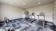 This dedicated exercise room at 522 Sweetgrass Dr in Brookings, South Dakota, offers a clean, modern fitness environment designed for effective cardio workouts. The space is thoughtfully outfitted with multiple treadmills, an elliptical trainer, and a rowing machine, providing a well-rounded selection of cardiovascular equipment for residents or users of varying fitness levels.

The room features a contemporary aesthetic, with neutral gray walls that create a calm, focused atmosphere. The floor is covered in a striking geometric-patterned carpet tile in varying shades of gray, adding both visual interest and durability underfoot. Recessed lighting illuminates the space evenly, complemented by natural light pouring in through large windows along one wall.

These expansive windows are a standout feature, framing a broad, open view of the surrounding South Dakota landscape — lush greenery and wide-open fields that provide an inspiring backdrop during workouts. An accent wall with a bold circular patterned wallcovering adds a design-forward touch, enhancing the room's modern character.

The layout is efficient, making excellent use of the available space while keeping the room uncluttered and easy to navigate. Overall, this exercise room delivers a bright, well-appointed, and motivating environment for daily fitness routines.