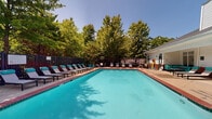 The Somerhill Farms Pool area in Gainesville, Virginia offers a well-appointed outdoor aquatic retreat designed for relaxation and recreation. At the heart of the space is a generously sized rectangular swimming pool with crystal-clear turquoise water, framed by a classic brick-accented surround and multiple entry points with stainless steel handrails. The expansive pool deck is lined with a plentiful arrangement of cushioned chaise lounges accented in teal, along with additional seating areas featuring outdoor tables and chairs — ideal for socializing or unwinding in the sun. A dedicated lounge nook adjacent to the clubhouse building provides a shaded seating area with a large sectional sofa in teal upholstery and a coffee table, offering a comfortable spot to relax out of direct sunlight. The pool area is enclosed by a dark metal fence, providing both security and an open feel, while mature trees surrounding the perimeter create a lush, green backdrop that enhances the sense of privacy. The on-site clubhouse building, finished in white siding with brick detailing, houses restroom facilities and is accessible directly from the pool deck via double glass doors. The entire space is bathed in abundant natural sunlight, making it a vibrant and inviting outdoor amenity.