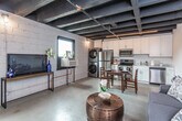 This single-floor urban loft masterfully combines industrial aesthetics with contemporary living. The space is defined by its striking exposed beam ceiling, painted in deep black, which runs throughout and lends an unmistakable raw, warehouse-inspired character. Polished concrete floors provide a seamless, cohesive foundation across all areas of the unit.

The open-plan living area comfortably accommodates a sitting space and a sleeping area within the same expansive room, separated naturally by layout rather than walls. The living zone features a grey loveseat and a distinctive copper-toned drum coffee table, while the sleeping area is anchored by a tufted blue velvet headboard and a large window that draws in natural light and frames views of the surrounding brick exterior.

The kitchen is fully equipped with stainless steel appliances, including a refrigerator, electric range, built-in microwave, and dishwasher. White shaker-style cabinetry provides ample storage, and a stacked washer and dryer unit is conveniently integrated into the kitchen space. A small dining table with chairs sits adjacent to the kitchen, offering a dedicated dining area.

The bathroom is cleanly finished with white cabinetry, a quartz-style countertop vanity, and a full tub-shower combination. Exposed concrete block walls in select areas reinforce the loft's industrial identity, creating a thoughtfully balanced blend of utilitarian design and modern comfort.