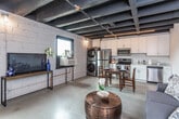 This single-floor urban loft masterfully combines industrial aesthetics with contemporary living. The space is defined by its striking exposed beam ceiling, painted in deep black, which runs throughout and lends an unmistakable raw, warehouse-inspired character. Polished concrete floors provide a seamless, cohesive foundation across all areas of the unit.

The open-plan living area comfortably accommodates a sitting space and a sleeping area within the same expansive room, separated naturally by layout rather than walls. The living zone features a grey loveseat and a distinctive copper-toned drum coffee table, while the sleeping area is anchored by a tufted blue velvet headboard and a large window that draws in natural light and frames views of the surrounding brick exterior.

The kitchen is fully equipped with stainless steel appliances, including a refrigerator, electric range, built-in microwave, and dishwasher. White shaker-style cabinetry provides ample storage, and a stacked washer and dryer unit is conveniently integrated into the kitchen space. A small dining table with chairs sits adjacent to the kitchen, offering a dedicated dining area.

The bathroom is cleanly finished with white cabinetry, a quartz-style countertop vanity, and a full tub-shower combination. Exposed concrete block walls in select areas reinforce the loft's industrial identity, creating a thoughtfully balanced blend of utilitarian design and modern comfort.