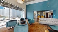 This single-floor space showcases a thoughtfully designed open-plan layout that seamlessly connects multiple functional zones, creating a cohesive and inviting environment. The design is defined by a striking teal shiplap accent wall that serves as a bold architectural centerpiece, visually anchoring the space while guiding movement between areas.

The lounge area features soaring double-height ceilings and expansive floor-to-ceiling windows that flood the interior with natural light and provide views of the surrounding outdoor environment. Seating arrangements include a mix of tufted sofas, mustard-yellow armchairs, and teal swivel chairs, all positioned atop patterned area rugs over rich hardwood flooring. A wall-mounted flat-screen television is integrated into the lounge area, adding a media focal point.

Adjacent to the lounge, a generously sized kitchen and bar area features a long dark-countertop island with built-in lighting along its base, accompanied by a row of sleek modern barstools. Dark wood cabinetry lines the back wall alongside open shelving and stainless steel appliances, including a full-size refrigerator. Recessed lighting throughout ensures consistent illumination across all zones.

A secondary lounge area with a darker ceiling and pendant lighting provides a more intimate atmosphere, complete with additional seating and outdoor access, adding versatility to the overall space.