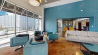 This single-floor space showcases a thoughtfully designed open-plan layout that seamlessly connects multiple functional zones, creating a cohesive and inviting environment. The design is defined by a striking teal shiplap accent wall that serves as a bold architectural centerpiece, visually anchoring the space while guiding movement between areas.

The lounge area features soaring double-height ceilings and expansive floor-to-ceiling windows that flood the interior with natural light and provide views of the surrounding outdoor environment. Seating arrangements include a mix of tufted sofas, mustard-yellow armchairs, and teal swivel chairs, all positioned atop patterned area rugs over rich hardwood flooring. A wall-mounted flat-screen television is integrated into the lounge area, adding a media focal point.

Adjacent to the lounge, a generously sized kitchen and bar area features a long dark-countertop island with built-in lighting along its base, accompanied by a row of sleek modern barstools. Dark wood cabinetry lines the back wall alongside open shelving and stainless steel appliances, including a full-size refrigerator. Recessed lighting throughout ensures consistent illumination across all zones.

A secondary lounge area with a darker ceiling and pendant lighting provides a more intimate atmosphere, complete with additional seating and outdoor access, adding versatility to the overall space.