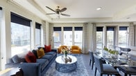 This single-floor space showcases a thoughtfully designed open-concept living and dining area that exudes contemporary elegance. The living room is anchored by a plush navy blue sofa accented with rich red and gold throw pillows, complemented by a pair of warm cognac leather accent chairs that add a bold pop of color. A round sculptural coffee table with a textured base sits atop a soft, shaggy area rug, adding layers of texture to the space.

Large windows along multiple walls flood the interior with natural light, dressed with a combination of navy blackout roller shades and patterned sheer curtains for versatile light control. The neutral-toned walls and light hardwood flooring provide a clean, cohesive backdrop that ties the design together. A ceiling fan with a brushed brass finish adds both function and a refined detail overhead.

The dining area transitions seamlessly from the living space, featuring a sleek table surrounded by upholstered chairs with nailhead trim detailing. Together, the two areas create a balanced, inviting environment well-suited for both relaxing and entertaining.