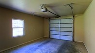 This single-story garage offers a straightforward and functional layout well-suited for vehicle storage or versatile use. The space is enclosed by smooth, tan-painted walls that lend a clean, neutral appearance throughout. A large paneled garage door dominates one wall, equipped with an automatic opener mounted to the ceiling via a sturdy rail system, providing convenient motorized access. The concrete floor spans the full footprint of the garage, offering a durable and easy-to-maintain surface appropriate for a range of uses beyond vehicle parking, including storage or a workshop setup. A single window positioned on the side wall introduces natural light into the interior, brightening the space without compromising wall area. The ceiling height is adequate for standard vehicles and overhead storage solutions. Overall, the garage presents a practical, no-frills environment that is move-in ready and adaptable to various needs.