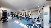 Located at 5901 John F. Kennedy Blvd in North Little Rock, Arkansas, this well-equipped fitness center offers a comprehensive workout environment designed to support a wide range of exercise routines. The single-floor space features durable blue rubber flooring throughout, providing both safety and comfort underfoot during workouts.

The cardio zone is prominently featured and includes multiple treadmills, elliptical trainers, stationary bikes, and an Assault Fitness rowing machine, offering users a variety of aerobic training options. A dedicated strength training area houses a multi-station cable machine, weight benches, and a dumbbell rack stocked with free weights, catering to resistance and functional training needs.

Full-length mirrored walls line one side of the room, visually expanding the space and allowing users to monitor their form. Wall-mounted flat-screen televisions provide entertainment during workouts. Recessed lighting and ceiling fans ensure the space remains bright and well-ventilated, while a wall-mounted air conditioning unit offers additional climate control.

Large sliding glass windows along one wall invite abundant natural light and offer views of the surrounding outdoor area, creating an open and energizing atmosphere. The overall design is clean, functional, and efficiently organized to maximize the available floor space for an effective fitness experience.