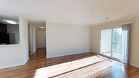 This single-floor space offers a clean, open layout highlighted by rich hardwood flooring that extends throughout the main living area and into the entryway corridor. The flooring transitions subtly in tone, adding visual depth and warmth to the interior. Neutral, light beige walls provide a versatile backdrop, complementing the white baseboards and ceiling trim that frame each surface crisply.

A sliding glass door along one wall serves as a prominent architectural feature, allowing abundant natural light to pour into the living space and creating a bright, airy atmosphere. The natural light casts defined beams across the floor, further accentuating the quality of the hardwood.

A hallway visible toward the rear of the space leads to the unit's entry door, which is fitted with standard hardware and flanked by a ceiling-mounted light fixture that provides functional illumination in that transitional zone. The corridor layout contributes a sense of depth to the overall floor plan.

The smooth, white ceiling runs at a consistent height throughout, maintaining a cohesive and uncluttered aesthetic. The overall ambiance is calm, neutral, and move-in ready, offering a functional foundation for a variety of interior arrangements.