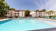 Located at 820 Macmillan SV in Wilmington, North Carolina, this outdoor amenity area centers around a large, resort-style swimming pool that serves as the focal point of the community. The crystal-clear pool is surrounded by a spacious concrete sundeck outfitted with bright blue lounge chairs and umbrella-shaded tables, providing ample space for relaxation and recreation under the Carolina sun. The pool area is enclosed by a black iron fence, ensuring a secure and well-defined environment. Adjacent to the pool is a well-appointed poolhouse or clubhouse structure, characterized by its coastal-inspired design featuring light blue and white trim, floor-to-ceiling glass panel doors, and a covered walkway with decorative exposed beam detailing overhead. This architectural element adds both function and charm, offering shaded access along the pool's edge. Mature trees line the perimeter, providing natural shade and a lush, green backdrop that softens the hardscape. The multi-story residential buildings visible in the background feature balconies that overlook the pool area, reinforcing the community-centric layout of the property. Overall, the space exudes a clean, well-maintained, and inviting atmosphere suited for outdoor leisure.