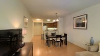 This single-floor residence offers an open-concept living and dining layout that maximizes the sense of space and promotes a natural flow between functional areas. The hardwood flooring runs throughout, lending warmth and continuity to the interior. The dining area is centrally positioned and accommodates a dark-finished rectangular table surrounded by cushioned chairs, complemented by elegant pendant lighting suspended from the ceiling above. Framed artwork adorns the walls, adding a refined touch to the space.

The kitchen is seamlessly integrated into the open layout via a breakfast bar peninsula, offering both casual seating and a practical workspace. Dark wood cabinetry lines the upper and lower walls of the kitchen, contrasting with lighter countertops. Standard appliances, including a refrigerator and microwave, are visible and well-positioned within the cabinetry arrangement. Recessed and flush-mount ceiling lighting provides even illumination throughout. The entry area is neatly defined, with a door featuring a sidelight panel, maintaining a clear separation between the unit's interior and the hallway beyond.
