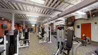 This fitness center combines industrial architectural character with a comprehensive array of modern exercise equipment. The space is defined by its striking exposed brick walls, painted structural columns, and an open ceiling that reveals white-painted steel trusses and mechanical systems overhead, lending the room a loft-like, urban aesthetic.

The floor is covered in a durable, multi-toned carpet tile, providing both comfort and practicality for a high-traffic fitness environment. Bright fluorescent lighting suspended from the ceiling trusses ensures the entire area is well-lit throughout.

The equipment layout is thoughtfully organized, with strength training machines — including cable stations, chest press, and isolation machines — positioned alongside a dedicated row of cardio equipment such as treadmills and ellipticals. Wall-mounted flat-screen televisions are strategically placed to be visible from multiple workout stations, enhancing the user experience during cardio sessions.

The open floor plan allows for generous spacing between machines, supporting a comfortable and safe workout environment. The blend of exposed brick, structural columns, and modern fitness equipment creates a distinctive atmosphere that balances raw industrial design with functional, contemporary fitness facility standards.