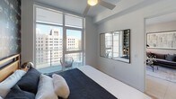 Located in the heart of Downtown Los Angeles, this contemporary residence at 825 S Hill St offers a refined urban living experience with sleek modern design throughout. The open-concept living area is anchored by expansive floor-to-ceiling windows and sliding glass doors that flood the space with natural light and frame sweeping views of the surrounding Downtown LA skyline. A private balcony extends the living space outdoors, providing a compelling vantage point over the city. The living room features stylish décor, a wall-mounted television, and curated artwork that complements the space's clean aesthetic. The kitchen is thoughtfully designed with a blend of warm wood cabinetry and crisp white upper cabinets, complemented by a generous island with seating — ideal for casual dining or entertaining. Stainless steel appliances and recessed lighting complete the functional yet refined kitchen. The bedroom continues the floor-to-ceiling window theme, bathing the room in natural light while offering direct balcony access and iconic city views. A ceiling fan adds comfort to the serene retreat. The bathroom is finished with large-format neutral stone tiles, a soaking tub with an overhead shower, and clean white fixtures, creating a spa-like atmosphere. Overall, the space exudes a sophisticated, contemporary ambiance rooted in its dynamic Downtown Los Angeles setting.
