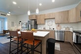 This thoughtfully laid-out one-bedroom, two-bathroom residence in Idaho Falls, Idaho, offers a clean, modern aesthetic throughout its single-floor layout. The kitchen is a standout feature, showcasing light wood cabinetry extending to the ceiling, bright white quartz countertops, stainless steel appliances including a range, dishwasher, and refrigerator, and stylish pendant lighting above a central island with bar seating — seamlessly connecting to the living area. Wood-look plank flooring runs throughout the main living spaces, adding warmth and continuity. The generously sized primary bedroom features plush carpet, a ceiling fan, a natural light-filled window with sheer curtains, and a warm, welcoming ambiance. Both bathrooms are finished with light wood vanities, white quartz countertops, wood-look flooring, and recessed lighting. The primary bathroom connects directly to the bedroom and includes a dual-sink vanity, providing added convenience. A hallway with panel doors offers access to additional storage or utility spaces. White walls and recessed lighting throughout contribute to a bright, airy feel that makes the space feel open and inviting. The overall design balances modern functionality with a cozy, neutral palette.
