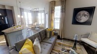This single-floor space showcases a thoughtfully designed open-concept layout that seamlessly connects the living, dining, and kitchen areas. The living room serves as the focal point, anchored by a tufted grey sectional sofa accented with bold yellow and patterned throw pillows. A geometric-patterned area rug and a sleek glass coffee table add modern sophistication to the space.

Large windows dressed with floor-length curtains in neutral tones allow generous amounts of natural light to flood the interior, creating an airy and welcoming atmosphere. The tall windows also offer views of the surrounding exterior, enhancing the sense of openness.

The adjoining dining area features a compact round table with modern seating, illuminated by contemporary track lighting overhead. The kitchen, partially visible from the living area, includes bar-height counter seating, reinforcing the open and social nature of the layout.

Dark hardwood-style flooring runs throughout, providing a warm contrast to the light-colored walls. The overall aesthetic is contemporary and cohesive, with the grey and yellow color scheme tying each zone of the space together harmoniously.
