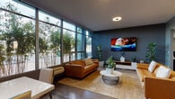 Located along the scenic Lake Merritt Path in Oakland, California, this single-floor space showcases a thoughtfully designed open-concept layout that seamlessly connects the living, dining, and kitchen areas. The interior is defined by striking floor-to-ceiling steel-framed windows that flood the space with natural light while offering lush views of mature bamboo plantings just outside, creating a tranquil, urban-garden atmosphere.

The living area features polished concrete floors throughout, dark blue-gray walls, and recessed lighting that contribute to a contemporary, sophisticated ambiance. A tan leather sofa anchors the lounge area, complemented by a round white coffee table, a wall-mounted flat-screen television, and a sleek media console. Potted plants are thoughtfully placed throughout, reinforcing the connection to the outdoors.

The dining area accommodates a white rectangular table with upholstered chairs, positioned adjacent to the expansive windows. Colorful abstract artwork punctuates the walls, adding visual interest and warmth. A glass partition provides a view into a secondary space beyond.

The kitchen is outfitted with stainless steel appliances, dark cabinetry, and a light-colored countertop with an integrated sink. Recessed ceiling lights illuminate the workspace effectively. The overall aesthetic balances industrial elements with modern comfort.