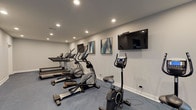 This fitness center is thoughtfully designed to support a full range of workout routines in a clean, modern environment. The space is organized across two connected zones, allowing for a natural separation between cardio and strength training areas. The cardio section features a row of treadmills, elliptical trainers, and stationary bikes, with a wall-mounted flat-screen television and decorative abstract artwork providing an engaging backdrop. Recessed ceiling lighting keeps the area well-lit and inviting.

The strength training zone is anchored by a Hoist multi-station cable machine and additional free-weight equipment, offering versatile options for resistance workouts. A bold teal accent wall adds a distinctive design element, contrasting with the neutral gray walls and crisp white trim throughout. Blue-toned carpet flooring runs across the entire space, providing comfort and visual cohesion.

Large windows allow ample natural light to fill the strength training area, creating an airy atmosphere. French doors with glass pane detailing connect sections of the facility, enhancing the open feel. Decorative touches such as potted plants and framed artwork contribute to a polished, welcoming ambiance. The space is well-maintained and equipped to serve the everyday fitness needs of its users.