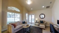 This single-floor business center offers a thoughtfully designed lounge area that balances comfort with functionality. The space is characterized by soaring vaulted ceilings that lend an open, airy feel, complemented by warm, neutral-toned walls that create an inviting atmosphere throughout.

Natural light floods the interior through a large arched window fitted with shutters, as well as a set of glass French doors that provide a seamless visual connection to the outdoors and allow ample sunlight to filter in. Recessed lighting and an elegant ceiling fixture with a decorative glass shade provide additional illumination for evening use.

The lounge area is furnished with plush, neutral-upholstered sofas and armchairs accented with teal and warm-toned throw pillows, arranged around an oval coffee table atop a rich, dark area rug. A coordinating ottoman and side table with a classic table lamp round out the seating arrangement, making the space well-suited for both casual conversation and focused work sessions.

A decorative round mirror with a textured dark frame serves as a tasteful focal point on the wall, adding depth and visual interest. The overall ambiance is warm, polished, and welcoming, making this business center an ideal setting for residents or guests seeking a comfortable and professional environment.