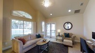 This single-floor business center offers a thoughtfully designed lounge area that balances comfort with functionality. The space is characterized by soaring vaulted ceilings that lend an open, airy feel, complemented by warm, neutral-toned walls that create an inviting atmosphere throughout.

Natural light floods the interior through a large arched window fitted with shutters, as well as a set of glass French doors that provide a seamless visual connection to the outdoors and allow ample sunlight to filter in. Recessed lighting and an elegant ceiling fixture with a decorative glass shade provide additional illumination for evening use.

The lounge area is furnished with plush, neutral-upholstered sofas and armchairs accented with teal and warm-toned throw pillows, arranged around an oval coffee table atop a rich, dark area rug. A coordinating ottoman and side table with a classic table lamp round out the seating arrangement, making the space well-suited for both casual conversation and focused work sessions.

A decorative round mirror with a textured dark frame serves as a tasteful focal point on the wall, adding depth and visual interest. The overall ambiance is warm, polished, and welcoming, making this business center an ideal setting for residents or guests seeking a comfortable and professional environment.