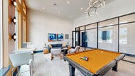 This dynamic game room in Scott, Louisiana, is thoughtfully designed to serve as a premier social and recreational hub. The space is defined by soaring ceilings that amplify its open, airy feel, complemented by light wood-style flooring that runs throughout. A bold, dual-toned color palette — rich amber-orange and deep teal accent walls — gives the room a lively, contemporary character.

At the center of the room stands a full-size pool table with a striking amber felt surface, serving as the room's focal point. Along the teal accent wall, a built-in cabinet system with dark navy cabinetry and marble-look countertops provides ample storage, topped by floating shelves and a mounted flat-screen television — ideal for entertainment. A sculptural, branch-style chandelier overhead adds an artistic touch to the space.

Floor-to-ceiling windows along the exterior wall flood the room with natural light while offering open views to the outdoors. The lounge area features a blue sofa, mid-century modern accent chairs, and upholstered ottomans arranged around a coffee table with a chess set, encouraging relaxed social interaction. Abstract artwork adorns the walls, reinforcing the room's modern aesthetic. Black-framed glass partition walls provide a sleek transition to adjacent areas, completing this well-appointed gathering space.