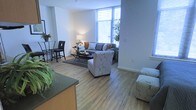 This single-floor residence offers a well-designed open-concept layout that seamlessly connects the kitchen, dining, and living areas into one cohesive living space. Warm hardwood-style flooring flows throughout, lending a clean and contemporary feel to the interior.

The living area is a standout feature, anchored by a large sectional sofa accented with colorful throw pillows and complemented by a patterned armchair and coordinating ottoman. A glass-top coffee table and area rug define the seating zone, while a tall floor lamp adds ambient lighting. The space is flooded with natural light through expansive floor-to-ceiling windows fitted with blinds, offering views of the surrounding urban landscape and bringing an airy, open atmosphere to the room.

Adjacent to the living area, a bistro-style dining set with sleek black chairs provides an intimate dining option. The kitchen, partially visible in the foreground, features dark countertops and light wood cabinetry, reflecting a modern design sensibility. Greenery is thoughtfully incorporated throughout, with potted plants adding life and vibrancy to the space.

The overall ambiance is modern, bright, and inviting — ideal for both relaxed everyday living and casual entertaining.