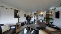 This single-floor residence offers an open, flowing layout that connects the main living area, dining space, and kitchen in a cohesive and functional arrangement. The living room serves as the central gathering space, featuring neutral-toned carpet flooring and white walls that create a bright, clean backdrop. The room accommodates a television and media console, complemented by decorative wall art and personal touches that give the space a lived-in warmth. A glass-top coffee table and seating anchor the lounge area, while decorative greenery adds a natural element throughout. Beyond the living space, a compact dining area provides room for a table and chairs, positioned near the kitchen entry where natural light filters through. The kitchen is visible in the background, featuring wood-toned cabinetry and overhead lighting. The open sightlines between rooms enhance the sense of space, making the layout feel connected and easy to navigate. Overall, the residence presents a comfortable, practical living environment with a casual and welcoming ambiance suited to everyday living.
