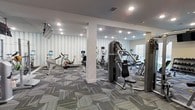 This single-floor fitness center offers a comprehensive workout environment designed to accommodate a variety of exercise routines. The space is outfitted with an array of cardio equipment, including stationary bikes and elliptical machines, each equipped with individual display screens for an interactive workout experience. Strength training machines are also present, providing users with options for resistance and muscle-building exercises.

The interior design balances functionality with aesthetics. A distinctive geometric-patterned wallcovering in soft blue and white tones lines the walls, adding visual interest without overwhelming the space. The flooring consists of a modern, multi-toned carpet tile in shades of gray and yellow, contributing both comfort underfoot and a contemporary look. Recessed lighting is evenly distributed across the white ceiling, ensuring the entire area is well-lit. A structural column near the center of the room subtly divides the space into functional zones. A window on one wall allows some natural light to enter. The overall atmosphere is clean, organized, and purposefully designed for regular fitness use.