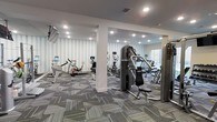 This single-floor fitness center offers a comprehensive workout environment designed to accommodate a variety of exercise routines. The space is outfitted with an array of cardio equipment, including stationary bikes and elliptical machines, each equipped with individual display screens for an interactive workout experience. Strength training machines are also present, providing users with options for resistance and muscle-building exercises.

The interior design balances functionality with aesthetics. A distinctive geometric-patterned wallcovering in soft blue and white tones lines the walls, adding visual interest without overwhelming the space. The flooring consists of a modern, multi-toned carpet tile in shades of gray and yellow, contributing both comfort underfoot and a contemporary look. Recessed lighting is evenly distributed across the white ceiling, ensuring the entire area is well-lit. A structural column near the center of the room subtly divides the space into functional zones. A window on one wall allows some natural light to enter. The overall atmosphere is clean, organized, and purposefully designed for regular fitness use.