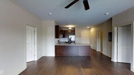 This single-floor space showcases a thoughtfully designed open-concept layout that seamlessly connects the living and kitchen areas. Dark hardwood flooring extends throughout, providing a warm and grounded aesthetic that contrasts elegantly with the light-toned walls and ceilings.

The kitchen is a standout feature, equipped with rich dark wood cabinetry that offers generous storage. A peninsula-style island with a dark wood base and light countertop serves as a natural dividing point between the kitchen and living areas, providing additional workspace and casual seating potential. Stainless steel appliances, including a refrigerator with an integrated water dispenser, add a modern, functional touch. A tile backsplash completes the kitchen's polished look.

Crown molding runs along the ceiling perimeter, adding a refined architectural detail throughout the space. Recessed lighting provides consistent illumination, supplemented by a ceiling fan in the living area for added comfort. Doorways visible along the hallway suggest multiple additional rooms, hinting at a well-organized floor plan. The overall ambiance is clean, modern, and functional.