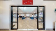 This well-appointed single-floor clubhouse offers a generous and thoughtfully designed communal space ideal for gathering, relaxing, and conducting business. The entrance greets visitors through floor-to-ceiling glass doors, immediately revealing an expansive open-plan interior bathed in natural light from large windows along the perimeter walls. Striking herringbone-patterned hardwood flooring flows consistently throughout, lending a polished, cohesive aesthetic to every area.

The central lounge area is furnished with plush navy blue sofas, accent chairs, and a rustic wood coffee table, all anchored by a patterned area rug. Textured accent walls and curated artwork contribute warmth and visual interest. Multiple seating configurations support both casual socializing and small gatherings. Large windows overlook outdoor spaces, enhancing the bright, airy atmosphere.

The kitchen is fully equipped with light maple cabinetry, granite-style countertops, stainless steel appliances including a full-size refrigerator and double wall oven, and a blue accent backsplash, making it well-suited for catering and events.

Separate private rooms include a relaxed lounge with sectional seating and entertainment features, as well as a glass-door office or conference space. A staffed reception desk near the entrance completes this polished, multifunctional facility.