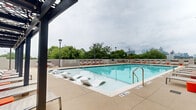 Located at 1777 Peachtree St NW in Atlanta's vibrant Midtown neighborhood, this outdoor amenity space centers around a generously sized resort-style swimming pool with crystal-clear turquoise water. The pool deck is thoughtfully designed with an expansive concrete surround that accommodates rows of cushioned chaise lounges and in-water sun loungers, providing ample space for relaxation and recreation. A pergola-covered lounge area along one side of the deck offers shaded seating, ideal for socializing or unwinding out of direct sun. Tall pole lighting is positioned throughout the deck, extending the usability of the space into the evening hours. The pool area is bordered by mature trees and low perimeter walls, creating a sense of privacy while still allowing for open sightlines. Notably, the deck offers impressive views of the Atlanta city skyline, particularly visible from the outer edges of the pool area. The modern high-rise building adjacent to the pool features multiple balconied residential floors with floor-to-ceiling windows, contributing to the contemporary architectural aesthetic of the overall space. Accent colors of orange and red on seating accessories add a vibrant pop against the neutral concrete and glass surroundings, reinforcing the lively, urban atmosphere of this well-appointed outdoor retreat.