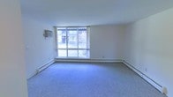 This single-floor residential unit offers a clean, functional layout designed for comfortable everyday living. The main living area is a standout feature, characterized by soft blue-grey wall-to-wall carpeting that adds warmth underfoot and a sense of continuity throughout the space. A large sliding glass door floods the room with natural light, creating an open and airy atmosphere while providing direct views of the outdoor courtyard and surrounding building exterior. A baseboard heating system runs along the perimeter of the living area, ensuring efficient and even warmth throughout the colder months, while a wall-mounted air conditioning unit provides climate control during warmer seasons. The walls are finished in a clean, neutral white, offering a versatile backdrop that complements a variety of interior styles. The overall ambiance of the unit is bright, understated, and move-in ready, with a straightforward design that prioritizes practicality. The connection to the outdoors through the generous glazing gives the living space a welcoming openness, making it feel larger than its footprint suggests.