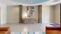 The lobby at West End Village, located at 3111 Long Blvd in Nashville, Tennessee, presents a thoughtfully designed communal entrance space that sets a polished, welcoming tone. The single-floor layout is anchored by large-format light tile flooring that runs throughout, lending a clean and open feel to the space.

A standout architectural feature is the curved tray ceiling finished in a soft blue-gray tone, which adds visual depth and a refined focal point overhead. Recessed lighting is distributed evenly across the ceiling, ensuring well-lit conditions throughout the day. Black-framed glass double doors at the entry allow natural light to filter into the lobby while offering views of the surrounding greenery outside.

The seating area is furnished with rich cognac leather armchairs and a sofa arranged on a layered area rug, creating an inviting lounge atmosphere. Flanking a central console table, a pair of bold decorative columns in a charcoal finish frame rotating curated artwork, adding a gallery-like quality to the space. Additional doorways lead to adjoining rooms, suggesting flexible utility beyond the primary lobby.

Overall, the space balances modern design with warm, residential comfort, making it an appealing communal hub for residents and visitors alike.