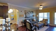 This single-story space showcases a well-appointed living area that blends comfort with thoughtful design. The room features rich hardwood flooring and elegant crown molding that frames the walls and ceiling, contributing to a polished, refined atmosphere. A decorative arched doorway connects the living area to an adjacent hallway, adding architectural interest and a sense of openness to the layout.

The living space is anchored by a plush gray sofa complemented by coordinating striped accent chairs and an upholstered ottoman, creating a cohesive and inviting seating arrangement. A large statement artwork depicting blossoming branches serves as a focal point on the main wall, flanked by matching teal ceramic table lamps that introduce a pop of color. A ceiling fan with a light fixture provides both illumination and practical comfort.

Natural light enters through a window fitted with blinds, offering views of the surrounding greenery outside. A decorative round mirror mounted near the arched entry adds depth and visual appeal to the space. The overall ambiance is warm and contemporary, striking a balance between stylish décor and functional, everyday living.
