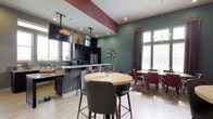 This single-floor space showcases a thoughtfully designed open-concept layout that seamlessly connects the kitchen, bar, and dining areas. The kitchen is equipped with dark cabinetry, stainless steel appliances, and a granite-topped island that doubles as a breakfast bar with contemporary bar stools. A ceiling-mounted television adds a functional touch above the kitchen area. The space is distinguished by bold accent colors, including a deep teal-green feature wall and a burgundy ceiling beam, creating a dynamic visual contrast against the light wood flooring. A round bistro-height table with leather-upholstered chairs serves as a central gathering point, while a separate dining area with burgundy and dark chairs provides additional seating. Large windows along the walls allow abundant natural light to fill the space, complemented by recessed lighting and a modern chandelier. Wall décor, including a sculptural metallic accent piece, adds artistic detail. The overall ambiance is modern and vibrant, balancing bold color choices with warm wood tones for an inviting, social atmosphere.