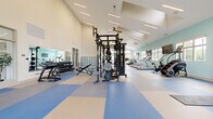 Located at 310 Northbend Drive in Charlotte, NC, this well-appointed fitness center offers a spacious, open layout designed to accommodate a wide variety of workout routines. The facility is housed within a single, generously sized floor featuring soaring vaulted ceilings with clerestory windows that flood the interior with natural light, creating a bright and energizing atmosphere throughout.

The flooring throughout the space consists of durable, color-blocked blue and cream interlocking tiles, providing both cushioning and visual zoning across different workout areas. The walls are finished in a calming seafoam green and warm white palette, contributing to an inviting and motivating environment.

The cardio zone is equipped with multiple treadmills, ellipticals, a stair climber, and rowing machines, complemented by wall-mounted flat-screen TVs. The strength training area features a multi-station cable machine, power racks, barbells, and a full dumbbell rack. A standout feature is the freestanding climbing wall — a "Freedom Climber" — offering a unique functional fitness option. Plyo boxes and stretching areas round out the functional training zones.

Large windows along the perimeter offer views of the surrounding community and landscaping, enhancing the sense of openness. A water station, built-in cubbies, and glass-panel entry doors connect the fitness center seamlessly to adjacent community amenities.