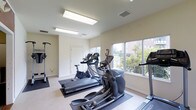 This fitness center offers a functional and straightforward workout environment designed to support a range of strength and conditioning exercises. The space is laid out on a single floor, providing an open and accessible area for users of all fitness levels.

The room is finished with neutral, off-white walls that contribute to a clean and focused atmosphere. Carpet-covered flooring in a muted gray tone adds both comfort underfoot and a degree of sound absorption, making the environment more conducive to exercise.

The fitness center is equipped with professional-grade machines, most notably a Cybex FT-325 functional trainer, a versatile cable-based unit capable of supporting a wide variety of upper body, core, and full-body strength exercises. Additional equipment is visible on the far side of the room, suggesting further options for resistance and strength training.

Overhead fluorescent lighting ensures the space is well-lit throughout, while ceiling-mounted HVAC vents indicate a climate-controlled environment to maintain comfort during workouts. The layout allows for adequate spacing between equipment, promoting safe and efficient use of the available floor space. Overall, this fitness center is a practical, purpose-built space suited for regular strength training and general exercise routines.