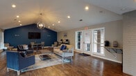 This single-floor community clubhouse is defined by its vaulted ceiling, which rises dramatically over the main gathering area and is highlighted by a stylish globe chandelier and recessed lighting. Rich hardwood flooring extends throughout the space, providing a warm, cohesive foundation across each distinct zone.

The main lounge area features a comfortable seating arrangement with a neutral tufted sofa, navy accent chairs, and a patterned area rug over a chrome-legged coffee table. A bold navy blue accent wall serves as the focal point, anchoring a wall-mounted flat-screen TV and flanking a dark wood media console. French doors and large windows along multiple walls flood the space with natural light and offer views of the surrounding brick exterior.

A dedicated workspace area near the back includes computer stations, while a separate sunroom-style lounge provides additional seating with abundant window coverage and direct outdoor access. A small granite-topped counter area with a coffee station is tucked near a painted brick column, adding a practical amenity to the welcoming environment. The overall ambiance is modern yet approachable, blending bold color accents with open, airy design.